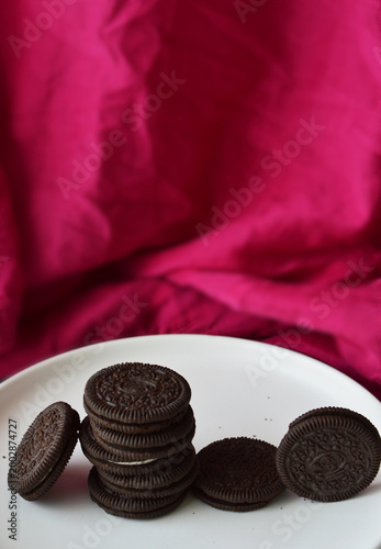 A stack of chocolate sandwich cookies is displayed on a white plate against a vibrant pink fabric backdrop.