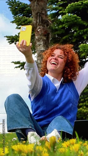 Happy student with red hair taking a selfie in a park while studying outdoors