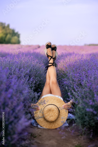woman lies in a field of lavender with her legs up. A braided hat and purple flowers around create a cheerful romantic mood.