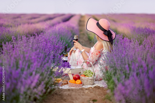woman in a big hat is sitting with a glass of red wine in a lavender field . Admiring the sunset in Provence. View from the back