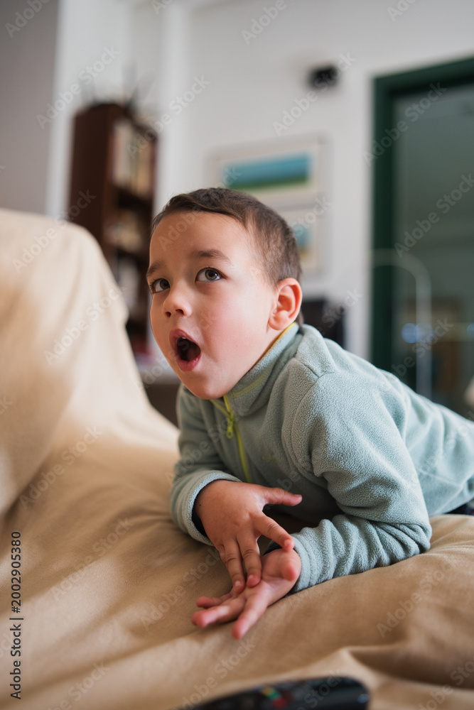 Fototapeta premium Young child lying on a couch with an open mouth, looking up in surprise or wonder while watching content on a screen in a domestic living room interior