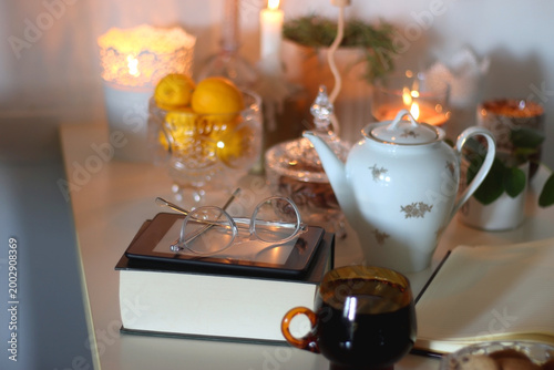 Cup of tea or coffee, teapot, book, notebook, e-reader, glasses, cookies, nuts, lemons, house plants and lit candles on the table. Hygge at home. Selective focus.