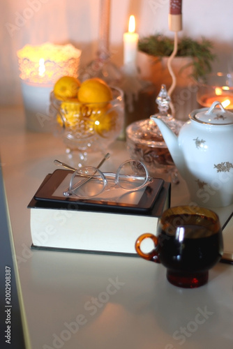 Cup of tea or coffee, teapot, book, notebook, e-reader, glasses, cookies, nuts, lemons, house plants and lit candles on the table. Hygge at home. Selective focus.