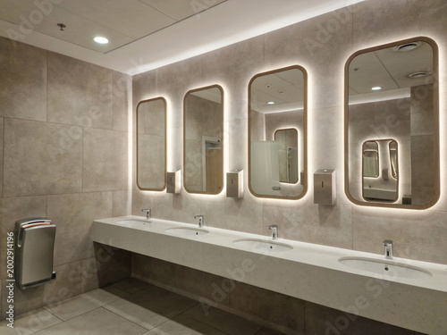 Modern public restroom with backlit mirrors, stone tiles, and white integrated vanity sinks