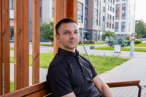 Man relaxing on wooden bench in city park. Young guy sitting outdoors in modern neighborhood. Rest and leisure time in urban environment. Calm daily life of student or office worker.