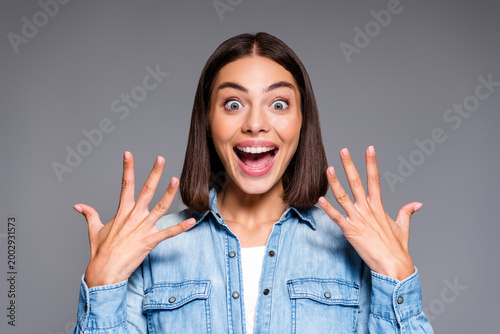 Young woman with excited surprised expression showing hands up in denim shirt, joyful portrait with wide eyes and open mouth on gray background
