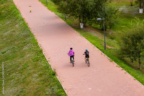 Woman and friend cycling on a paved path in a city park. Aerial view of athletic people riding bicycles outdoors. Concept of healthy lifestyle, active leisure and eco-friendly transport.