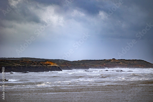 Konstfotografi Stormy coastal shoreline with waves crashing against rocky cliffs under dark clo