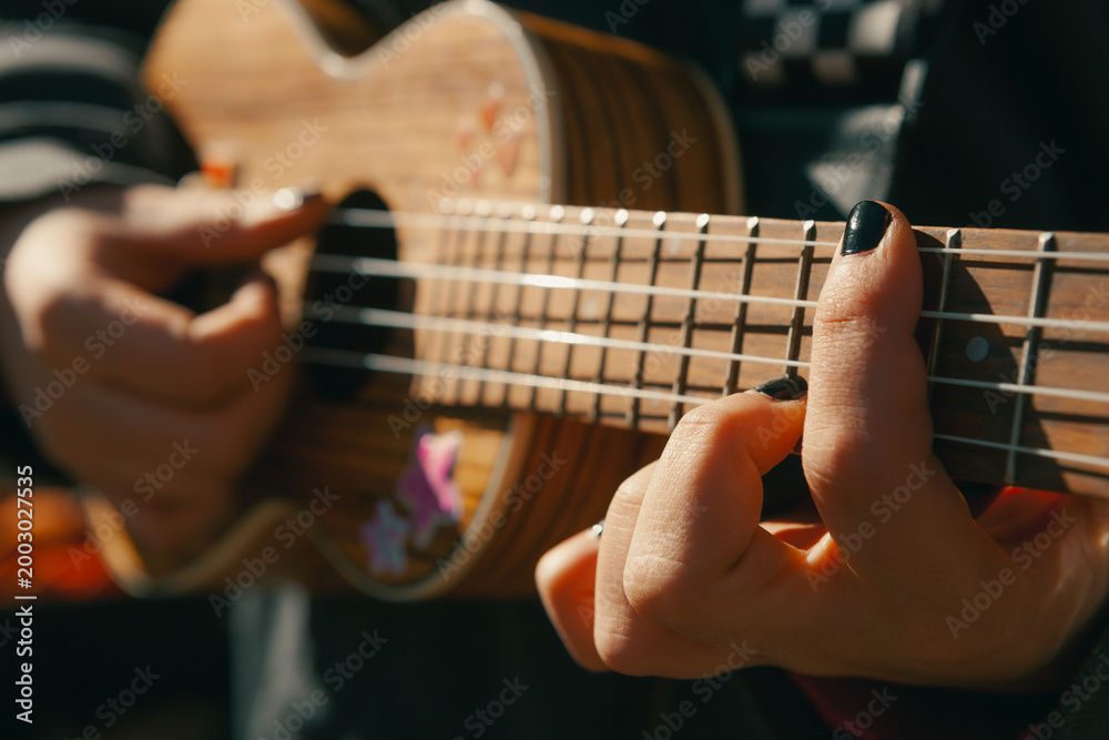 Fototapeta premium A close-up shot capturing the hands of a person with black nail polish playing a wooden ukulele adorned with star stickers, bathed in warm natural sunlight during an outdoor session.