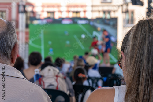 Fotografija People watching football match on large outdoor screen before game start at publ