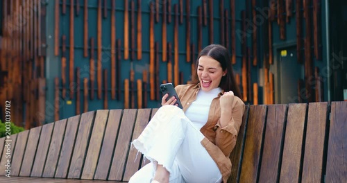 Smiling woman checking her smartphone and reacting to happy news outdoors, with a modern office building in the background.