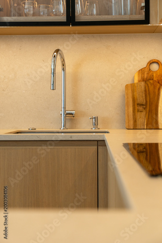 Fotografie A clean modern kitchen close-up showing a chrome gooseneck faucet over a stainless sink, warm stone countertop and wooden cutting boards leaning against a neutral backsplash for a minimalist look