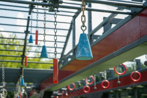 Red gymnastic rings hanging on chains in outdoor obstacle course in park, fitness and training equipment detail.