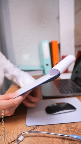 Vertical video medical professional working at a modern office desk, holding a clipboard with paper, reviewing healthcare documents, with a laptop, mouse pad, and stethoscope nearby