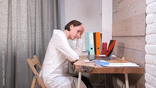Pensive female doctor wearing a white lab coat sitting at her desk, holding her forehead and contemplating work stress, healthcare challenges, and the demands of her profession in a clinic office