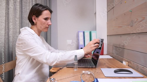 Woman in a lab coat spraying disinfectant cleaner on her laptop keyboard, maintaining hygiene and sanitation practices while working in a medical office environment