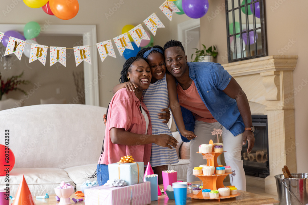 Obraz premium African American family hugging daughter in living room, celebrating with cupcakes, gifts, balloons