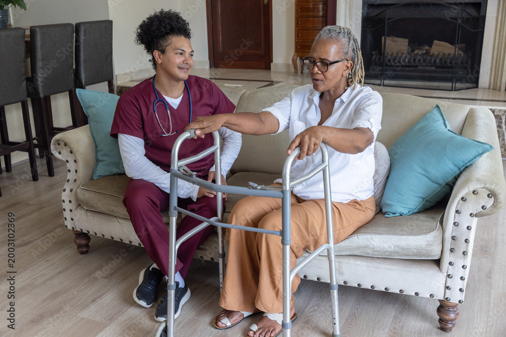 Fototapeta premium Senior African American woman sitting on sofa at home using walker, caregiver in scrubs watching