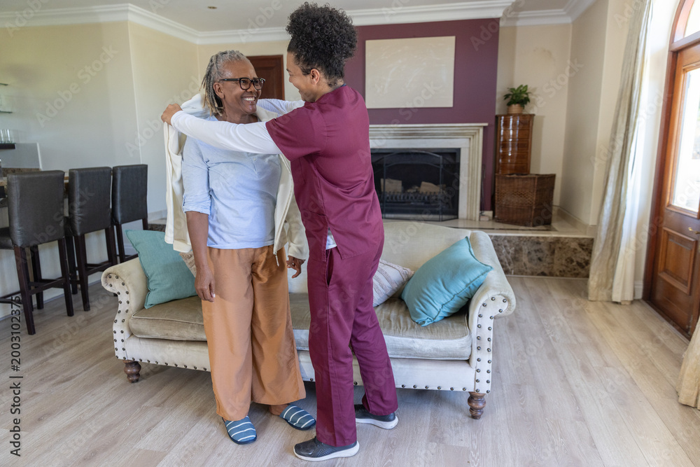 Fototapeta premium Mid-adult caregiver in maroon scrubs placing jacket on Senior African American woman in living room