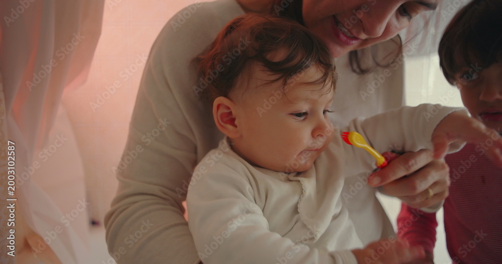 Naklejka premium Mother with baby and son in bathroom, smiling gently while helping infant explore toothbrush, warm family interaction showing affection, patience, and nurturing care