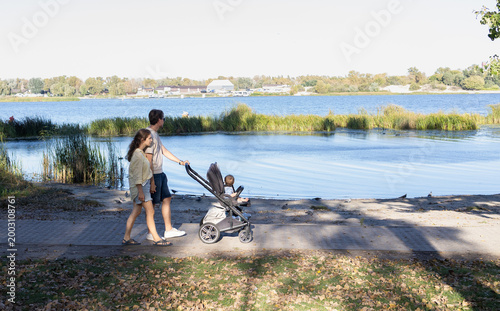 Bild auf Leinwand Young parents stroll with a stroller through a park next to a lake