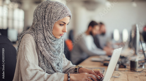 A young, modern Muslim woman wearing a hijab works on a laptop in a coworking space or office, typing on a laptop keyboard, looking at the screen. Businesswoman, computer, programmer, manager.