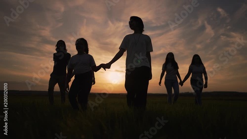 Large family group walk forward. Silhouette figures cross open grass field. Parent holds young child hand. Golden sunset brightens cloudy evening sky. Family silhouette walk towards warm sunset.