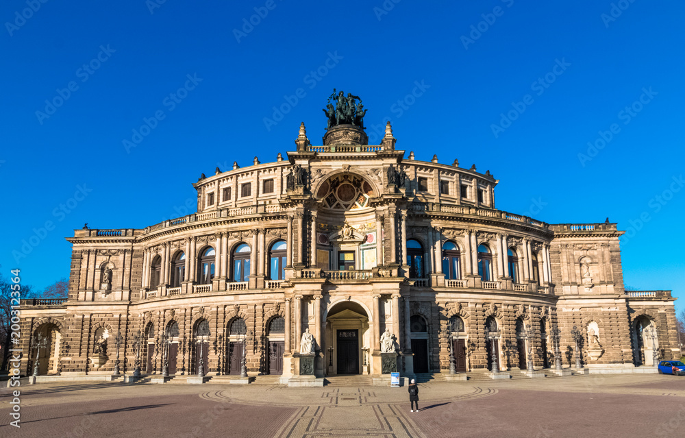 Obraz premium Picturesque front view of the Semperoper in Dresden, its sandstone Neo‑Renaissance façade illuminated by the sun, with the large central archway, deep semi‑circular exedra and sculptural decoration.