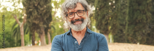 Retired man with glasses crossing his arms and looking at the camera with smile while standing in the park. Panoramic