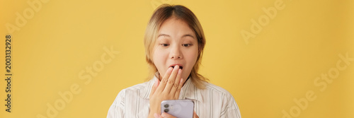Surprised young woman dressed in shirt and tie, with mobile phone in hands getting good news isolated on yellow background in studio. Panoramic