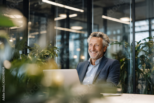 Smiling mature businessman in navy dotted blazer and open collar striped shirt, seated at laptop amid lush office plants and glass partitions
