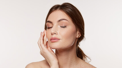 Woman with closed eyes touches cheek as fine white facial mesh covers face and neck on clean background. Ideal for skincare analysis, cosmetic treatments and beauty tech ads.