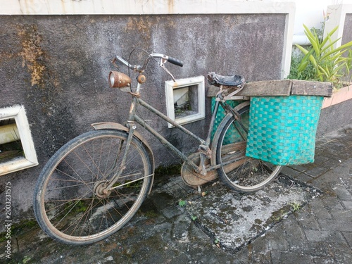 An old, rusty vintage bicycle with a torn saddle and large green woven bamboo baskets parked against a weathered concrete wall. The classic bicycle shows signs of age and heavy use.
