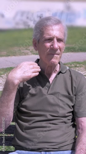 Vertical: Elderly man sitting on a park bench fans himself with his hand under extreme summer heat. The scene shows discomfort and the struggle to cope with high temperatures.