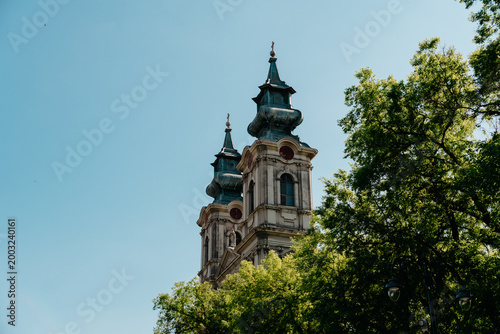 Cathedral of Saint Teresa of Avila rising above green trees in Subotica, Serbia. Baroque church towers stand against clear blue sky in historic city center.