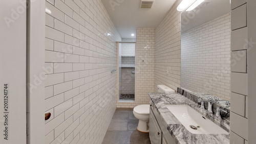 Narrow bathroom design with white subway tiles on walls and marble vanity top featuring chrome fixtures and grey stone floor leading to walk in shower area with built in shelving and neutral tones.