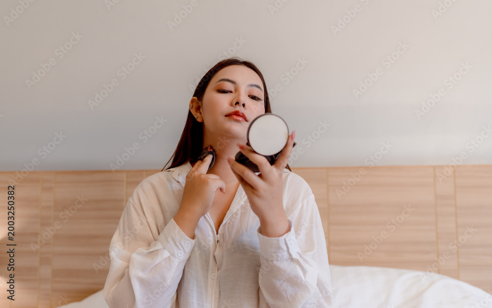 Φωτογραφία Young woman sitting on bed applying skincare or makeup while looking into a handheld mirror
