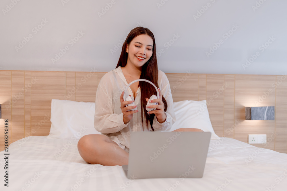 Φωτογραφία Young woman relaxing on bed while using a laptop and wearing headphones enjoying music or online content