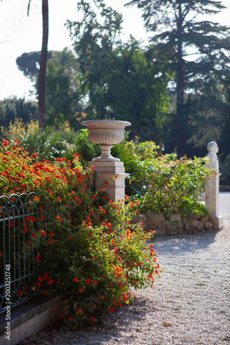 Ornate stone garden vase surrounded by lush green plants and flowers in a Roman park.