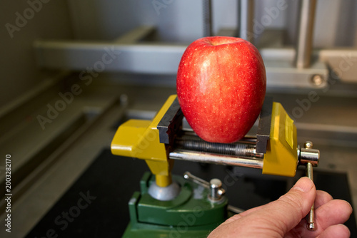 Red apple clamped in a metal vise held by a hand. Concept of food quality control, environmental pressure, or genetic engineering in agriculture.