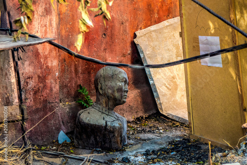 An old wooden sculptural head in an abandoned courtyard, surrounded by rubble, wooden panels and tangled cables. 