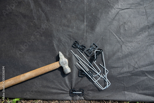 Fotografie A person wearing boots and gloves is kneeling on the ground on a flower bed installing black weed fabric using galvanized steel pegs and a mallet