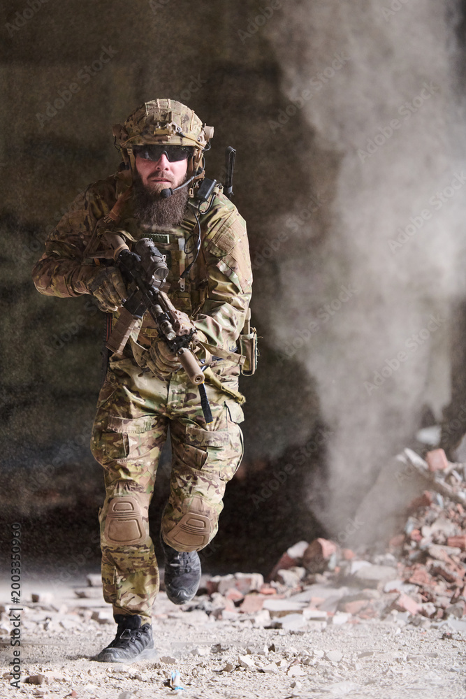 Fototapeta premium Bearded Special Forces Soldier Running Through Dusty Urban Ruins With Rifle And Tactical Gear