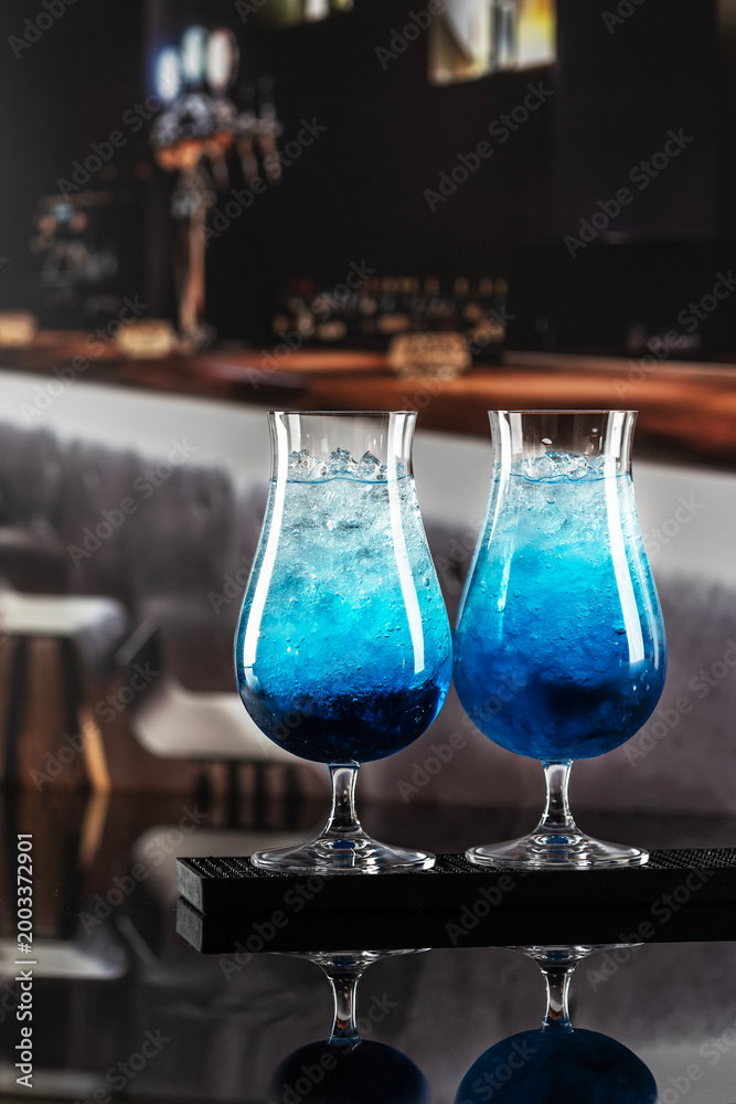 Fototapeta premium Close-up of colorful cocktail with blue Curacao in interior of restaurant or cafe. Refreshing drink with blue syrup. Blue lagoon drink with blue curacao at bar counter close up. Tall wine glasses