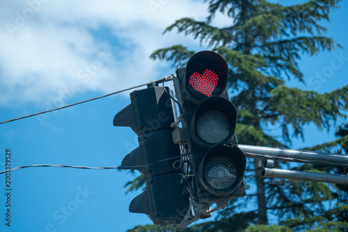 Traffic light with a glowing red heart signal against a blue sky and trees, symbolizing love, stop, road safety and urban street design.