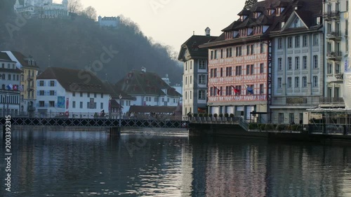 Local people walking across the historic Reussbrücke bridge, featuring traditional painted guild houses and Swiss flags along the riverfront in Lucerne.