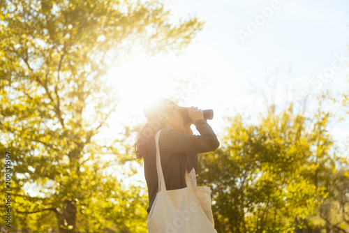 A young woman in a cap and jacket looks through binoculars during a bright, sunlit day outdoors, capturing a peaceful moment of nature exploration and wildlife observation.