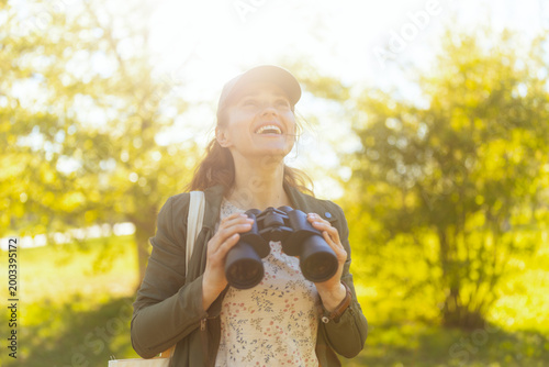 With a joyful and curious expression, a young woman holds her binoculars while looking up at the trees in a golden, sunlit park during a birding excursion.