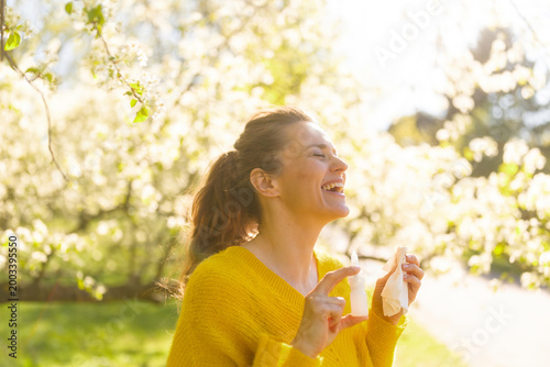 Canvas Print Smiling amidst blooming trees, a woman feels relief from seasonal allergies using a nasal spray and tissue, enjoying the fresh spring air in a sunny garden