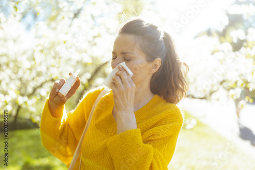 A woman deals with seasonal hay fever symptoms, using a tissue and nasal spray while standing in a beautiful but pollen-filled spring garden.